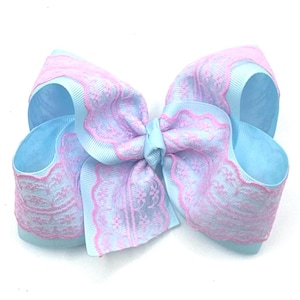 May include: A large, decorative hair bow in pastel colors. The bow features a light blue base with a layer of lavender lace and pink scalloped edges. The bow is tied in the center with a matching ribbon.