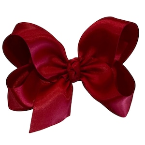 May include: A large, vibrant red satin bow. The bow is tied in the center and has multiple loops and tails. The fabric has a slight sheen, and the bow is set against a white background. This accessory is suitable for hair or gift wrapping.