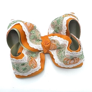 May include: A hair bow constructed from solid orange grosgrain ribbon layered with a white lace overlay featuring an embroidered pattern of orange pumpkins and green vines, finished with a central knot of matching orange ribbon.