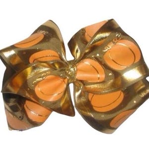 May include: A large, layered hair bow featuring a repeating pattern of orange pumpkins on a gold and brown background. The bow is tied in the center with a gold knot, creating a festive accessory.