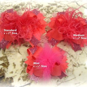 May include: Three pink and red hair bows with feathers. The bows are labeled "Standard 6 1/2" Size", "Medium 5 1/2" Size", and "Mini 4 1/2" Size".