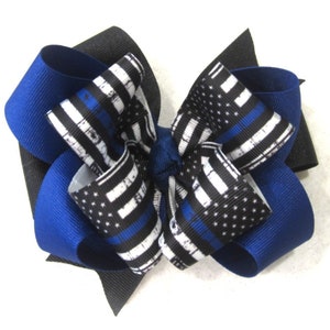 May include: A blue and black hair bow with a thin blue line American flag design. The bow is made of grosgrain ribbon and has a black center.