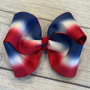 May include: Large, patriotic hair bow featuring a gradient of red, white, and blue. The bow is made of a wide, ribbed fabric and tied in the center. The bow is approximately 6 inches wide.