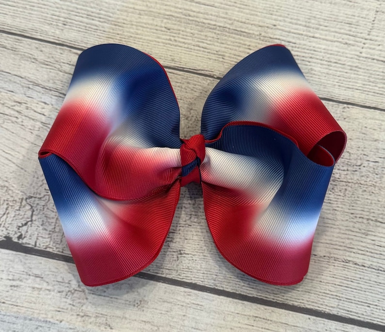 May include: Large, patriotic hair bow featuring a gradient of red, white, and blue. The bow is made of a wide, ribbed fabric and tied in the center. The bow is approximately 6 inches wide.