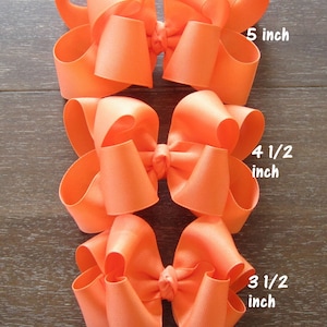 May include: Three orange hair bows of different sizes. The largest bow is 5 inches wide, the middle bow is 4 1/2 inches wide, and the smallest bow is 3 1/2 inches wide.