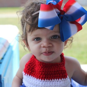 May include: A young child wearing a red, white, and blue crocheted halter top and a matching tutu. A large patriotic bow with red, white, and blue stripes adorns the child's hair. The outfit is perfect for a patriotic celebration.