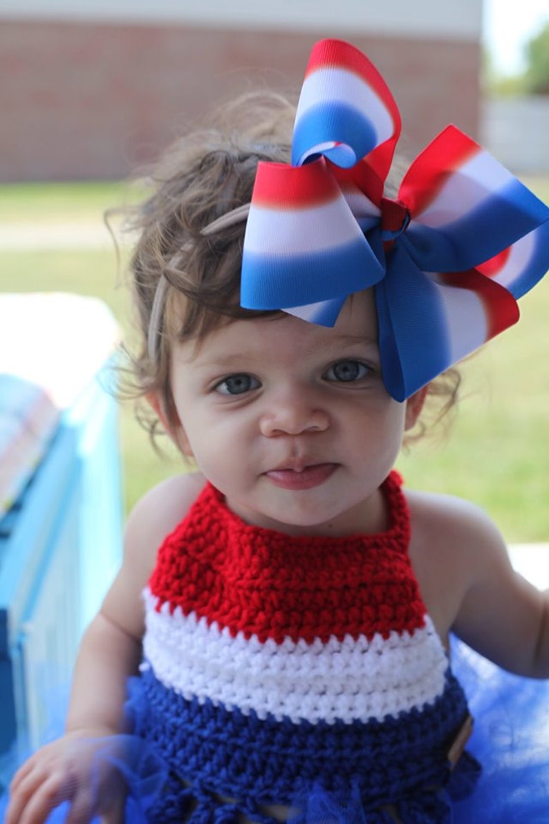 May include: A young child wearing a red, white, and blue crocheted halter top and a matching tutu. A large patriotic bow with red, white, and blue stripes adorns the child's hair. The outfit is perfect for a patriotic celebration.