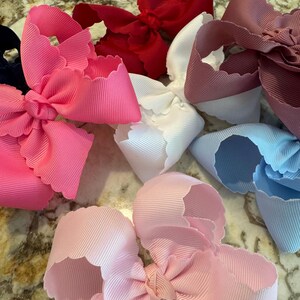 May include: A collection of six hair bows made from ribbed grosgrain ribbon with scalloped edges. The bows are featured in various solid colors including bright pink, deep red, white, dusty rose, light blue, and pale pink, each tied in a center knot.