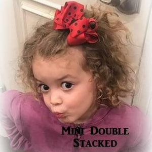 May include: A red and black polka dot hair bow with a double stacked design. The bow is worn by a young girl with curly hair. The text "Mini Double Stacked" is visible in the image.