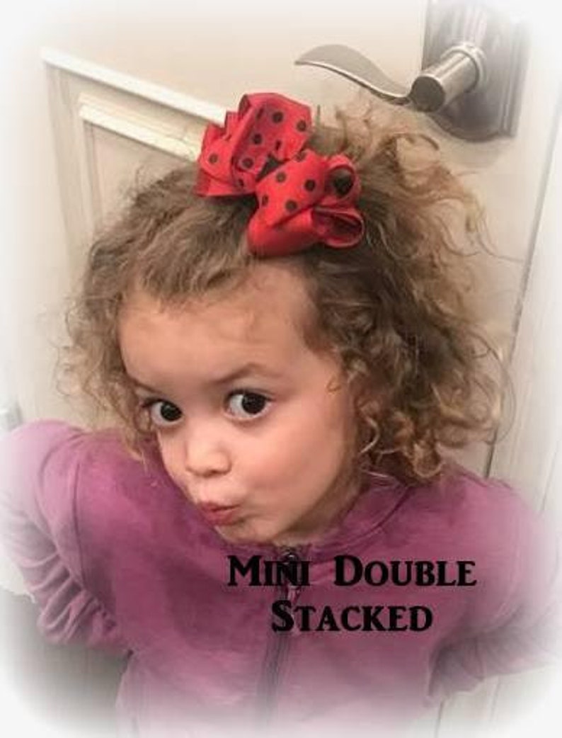 May include: A red and black polka dot hair bow with a double stacked design. The bow is worn by a young girl with curly hair. The text "Mini Double Stacked" is visible in the image.