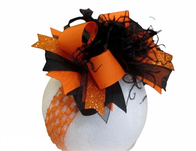 May include: A white, round Halloween decoration with orange and black accents. The decoration features an orange bow with glittery accents and black feathers. A decorative orange lace band wraps around the base of the white sphere.