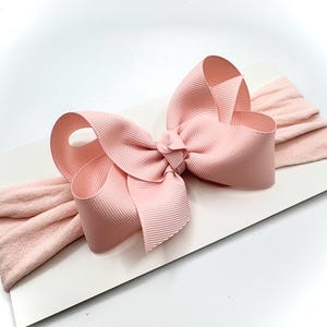 May include: A light pink baby headband featuring a large, textured bow. The bow is made of a wide, ribbed fabric and is centered on a soft, stretchy band. The headband is designed for infants and toddlers.