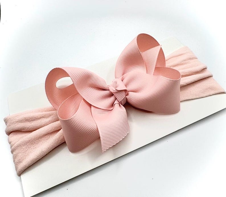 May include: A light pink baby headband featuring a large, textured bow. The bow is made of a wide, ribbed fabric and is centered on a soft, stretchy band. The headband is designed for infants and toddlers.
