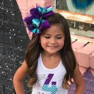 May include: A young girl with long brown hair wearing a white tank top with a purple and teal number four and the name "Harper" on it. She is wearing a large purple and teal bow with feathers in her hair.