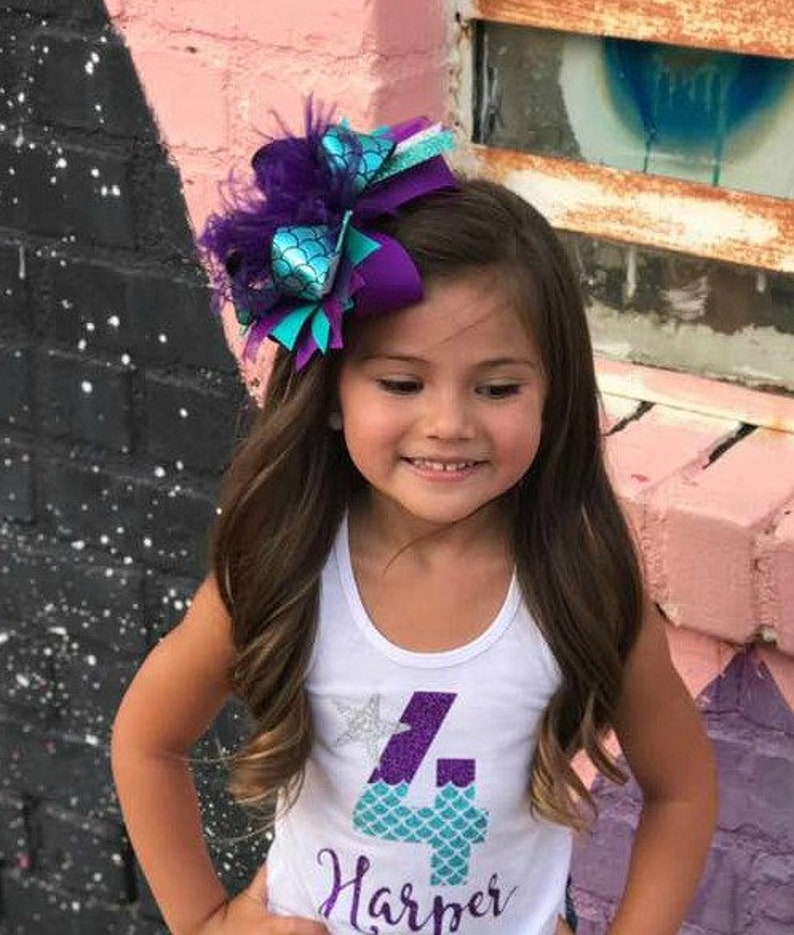 May include: A young girl with long brown hair wearing a white tank top with a purple and teal number four and the name "Harper" on it. She is wearing a large purple and teal bow with feathers in her hair.