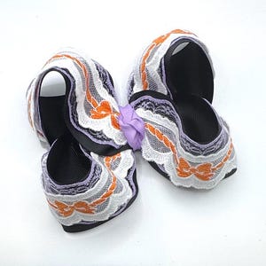May include: A hair bow constructed from black grosgrain ribbon layered with white lace trim featuring orange embroidered bow motifs and light purple accents. The center of the bow is gathered with a small, knotted light purple ribbon tie.