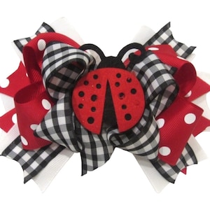 May include: A red and white polka dot ribbon, a black and white checkered ribbon, and a white ribbon are tied together in a bow with a red ladybug in the center.