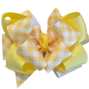 May include: A large, layered hair bow in shades of yellow and white. The bow features a gingham pattern on the top layer and solid yellow ribbon on the bottom layer. The bow is a cheerful accessory.