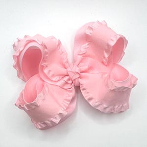 May include: A large, light pink hair bow with a ruffled edge. The bow is made of a soft, satin-like material and has a classic bow shape. The bow is centered and sits against a white background.