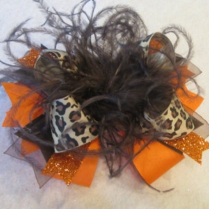 May include: A decorative hair bow with a mix of textures and colours. The bow incorporates orange, black, and leopard print ribbons, complemented by brown feathers and glitter. This accessory is designed to add flair to any hairstyle.