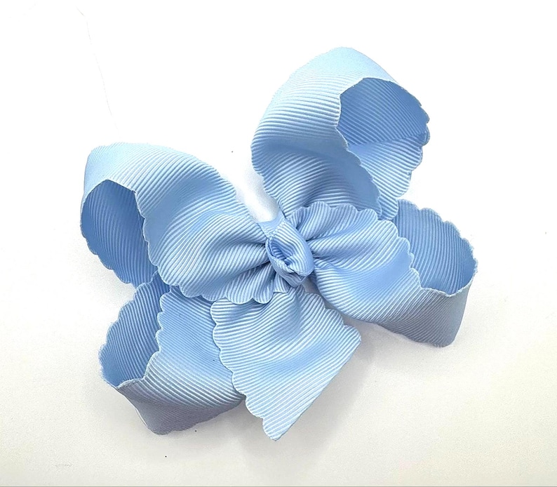 May include: A light blue hair bow constructed from ribbed grosgrain ribbon. The bow features a multi-layered design with scalloped edges along the perimeter of each loop, creating a voluminous and textured appearance against a plain white background.