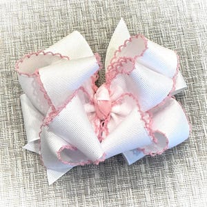 May include: A white hair bow with pink scalloped edges. The bow is made of a thick, textured ribbon and is tied in a large, fluffy knot.