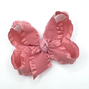 May include: A dusty rose hair bow featuring a textured fabric with ruffled edges. The center of the bow is gathered with a smooth, lighter pink ribbon knot, creating a layered, voluminous appearance with a soft, matte finish throughout the design.