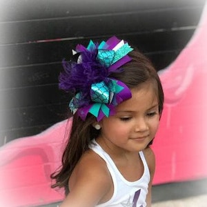 May include: A purple, teal, and white hair bow with a mermaid scale design and feathers. The bow is worn by a young girl with brown hair.