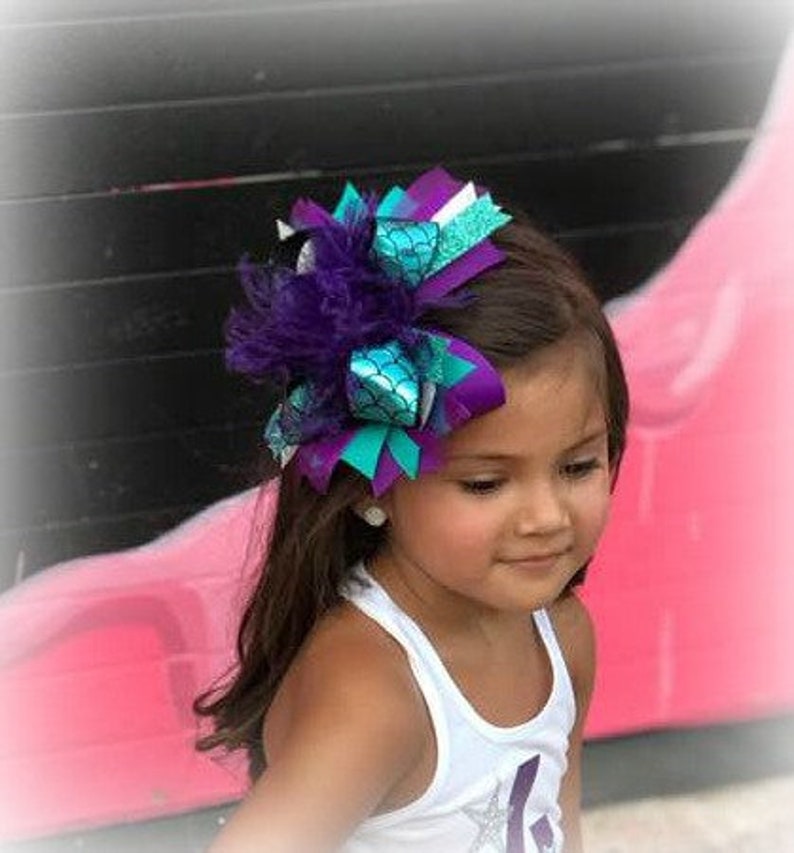 May include: A purple, teal, and white hair bow with a mermaid scale design and feathers. The bow is worn by a young girl with brown hair.