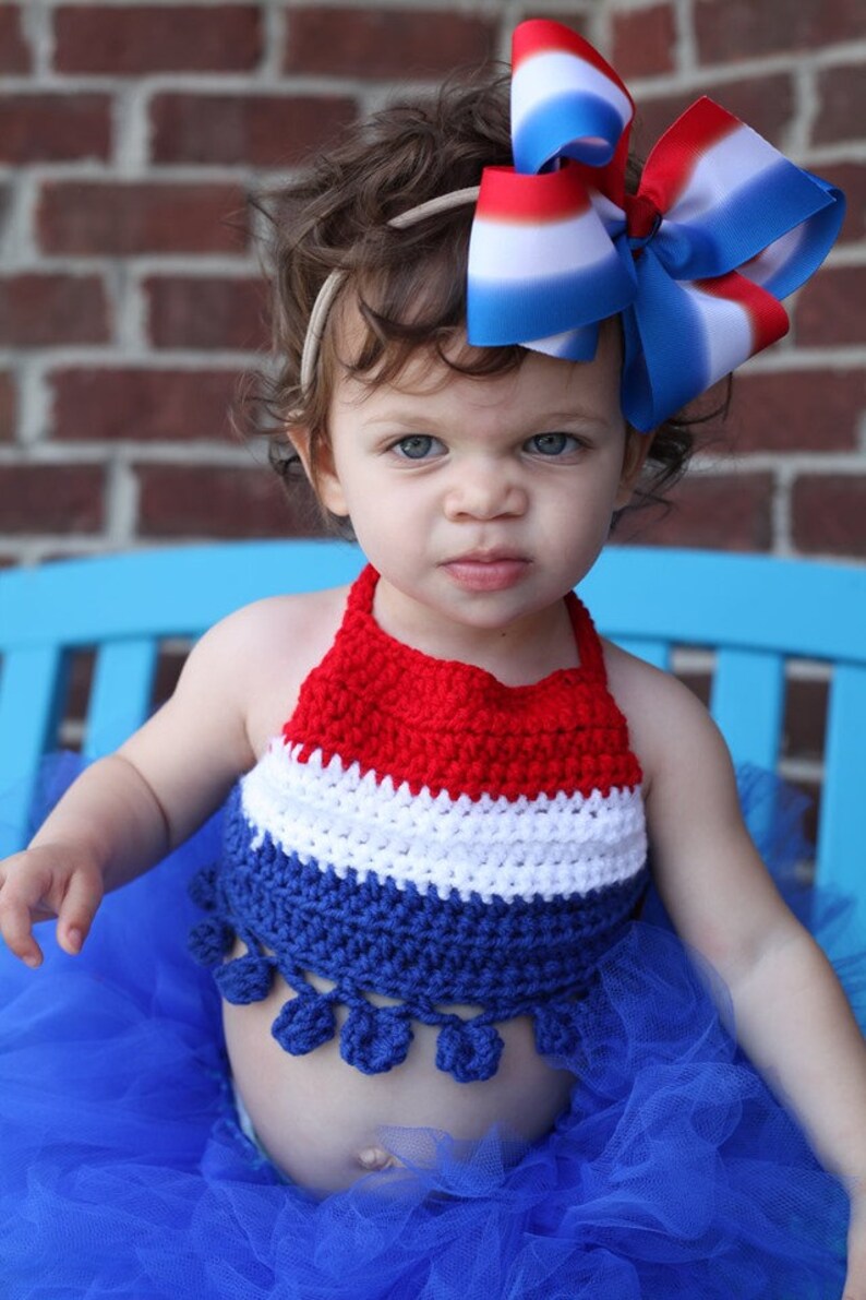 May include: A red, white, and blue crochet halter top and a blue tulle skirt. A large red, white, and blue bow is in the child's hair. The outfit is patriotic and suitable for a holiday.