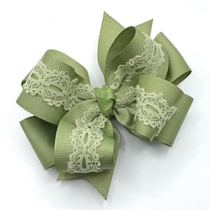 May include: A sage green hair bow with a layered design. The bow features a ribbed texture and is accented with delicate cream-coloured lace. The bow is suitable for various occasions and adds a touch of elegance.