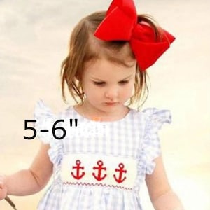 May include: A large red hair bow measuring 5-6 inches in width, paired with a light blue and white gingham dress featuring ruffled sleeves and a white smocked chest panel embroidered with three red anchors across the front of the garment.