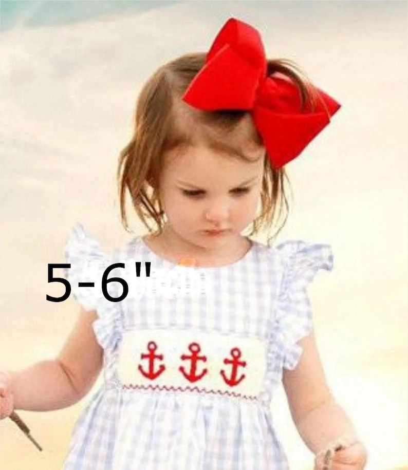May include: A large red hair bow measuring 5-6 inches in width, paired with a light blue and white gingham dress featuring ruffled sleeves and a white smocked chest panel embroidered with three red anchors across the front of the garment.