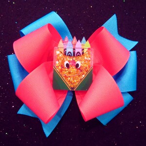 May include: A large, handmade hair bow featuring a vibrant color scheme. The bow has layers of bright pink and turquoise ribbon. At the center is a small, green box with a crayon design, filled with glitter and a smiling face.