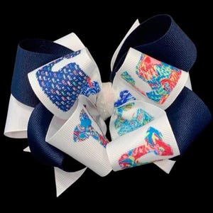 May include: A layered hair bow in white, navy blue, and patterned ribbon. The white ribbon has colourful, abstract designs. The bow is centered with a small, sparkly embellishment. The background is black.
