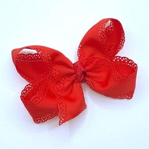 May include: A bright red hair bow with a decorative lace trim. The bow is made of a ribbed fabric and is tied in a classic bow shape. The lace trim adds a delicate touch to the accessory.