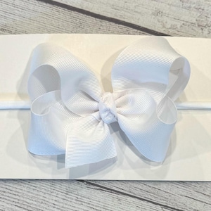 May include: White hair bow with a white elastic headband. The bow is made of a soft, textured fabric.