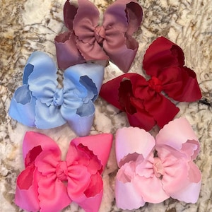 May include: Five hair bows made of ribbed fabric with scalloped edges. The collection includes one mauve, one light blue, one deep red, one bright pink, and one pale pink bow. Each bow features a central knot and layered, voluminous loops.