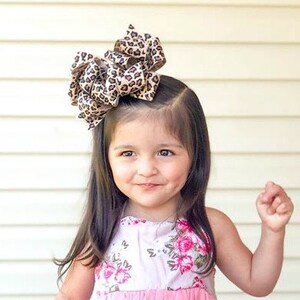May include: A young girl with long brown hair is wearing a pink and white floral dress. She has a large brown and black leopard print bow in her hair.