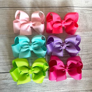 May include: Six large hair bows in various colors, including pink, light blue, purple, green, and red. The bows are made of a shiny, textured fabric and are tied with a knot in the center.