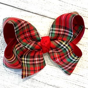 May include: A large, decorative bow featuring a classic red and green plaid pattern. The bow has a solid red center and edges, with a traditional tartan design in the main body. The bow is likely made of fabric and is suitable for various decorative purposes.