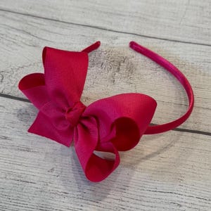May include: A bright pink satin bow headband. The headband is made of a thin, flexible metal and is adorned with a large, double-loop bow.