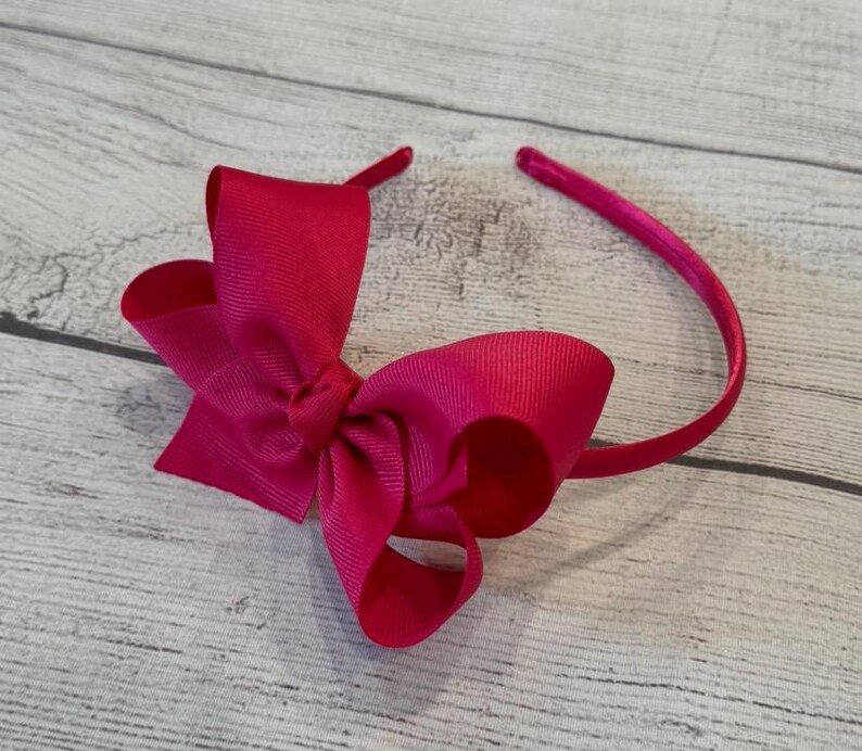 May include: A bright pink satin bow headband. The headband is made of a thin, flexible metal and is adorned with a large, double-loop bow.