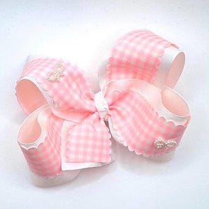 May include: A hair bow featuring a pink and white gingham pattern on a textured ribbon with scalloped edges. The bow is layered with solid white ribbon underneath and is accented with two small, silver-toned decorative bow charms.