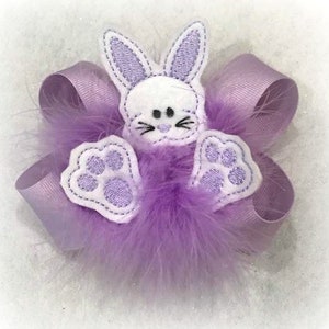May include: A purple and white bunny hair bow with a fluffy texture. The bunny has white fur, purple ears, and a white nose.