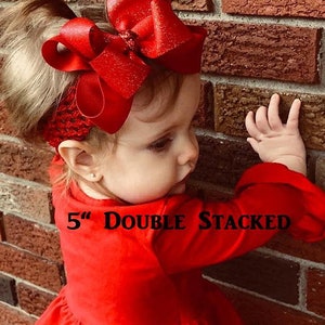 May include: A young girl wearing a red dress with a large, double-stacked red bow in her hair. The bow is 5 inches wide and has a sparkly finish. The text "5" Double Stacked" is visible on the dress.