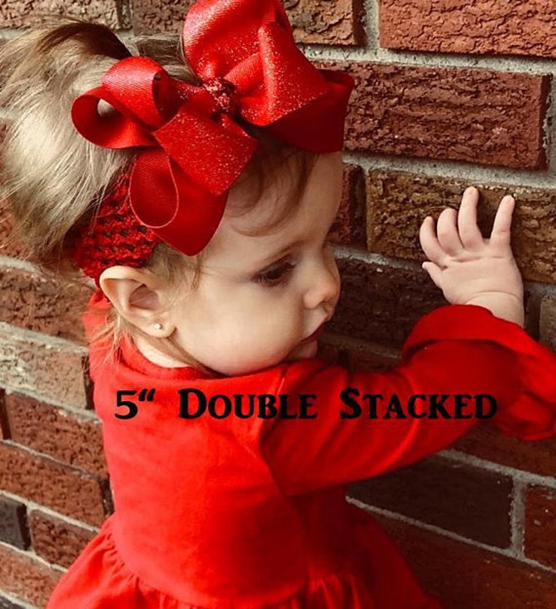 May include: A young girl wearing a red dress with a large, double-stacked red bow in her hair. The bow is 5 inches wide and has a sparkly finish. The text "5" Double Stacked" is visible on the dress.