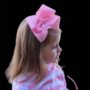 May include: A large, light pink bow hair accessory. The bow is made of a soft fabric and is attached to a child's head. The child is wearing a pink and white striped shirt. The background is black.
