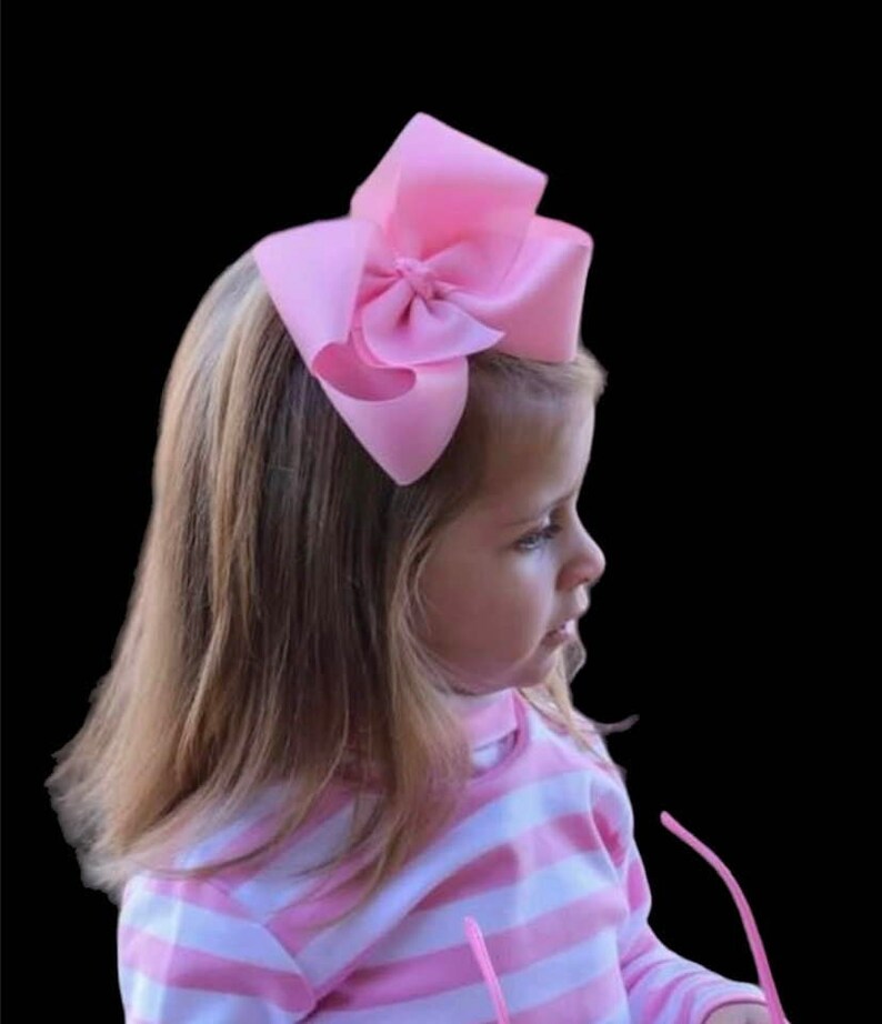 May include: A large, light pink bow hair accessory. The bow is made of a soft fabric and is attached to a child's head. The child is wearing a pink and white striped shirt. The background is black.