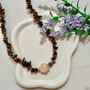 Puede incluir: Un collar que presenta cuentas de astillas de ojo de tigre en tonos marrón y dorado, acentuado por una cuenta central de piedra lisa en forma hexagonal de color melocotón claro, flanqueada por pequeñas cuentas espaciadoras de metal dorado.